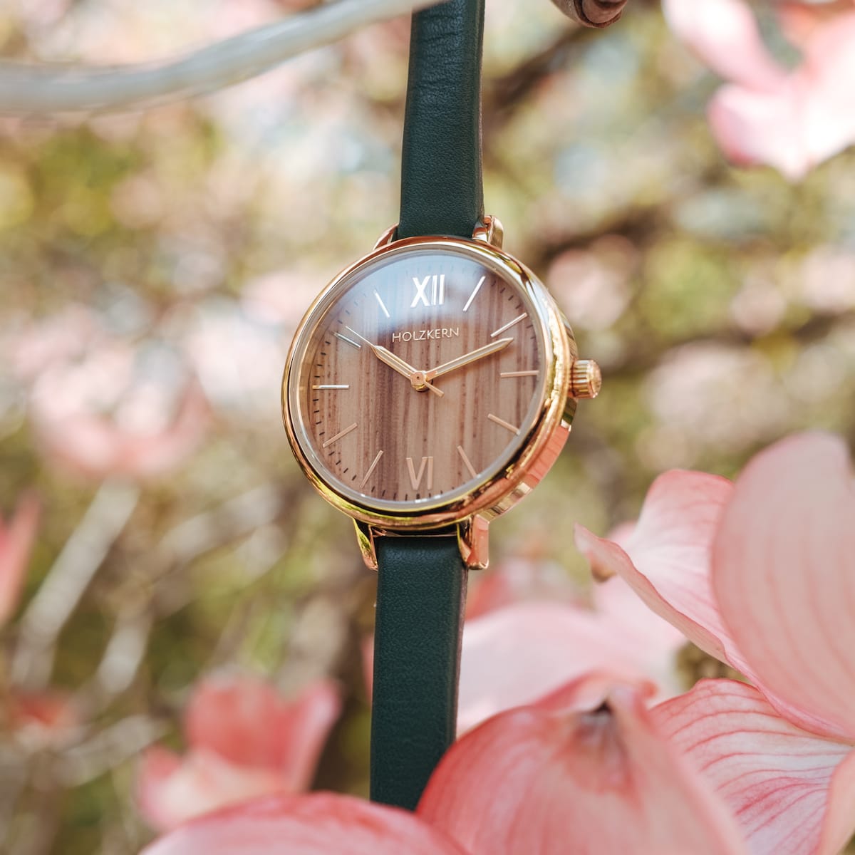 "Wangari" watch featuring zebrawood and gold accents, surrounded by pink petals and greenery.