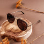 "The Honorable" sunglasses featuring a sleek, aviator style with leadwood and silver accents against a natural wood backdrop.