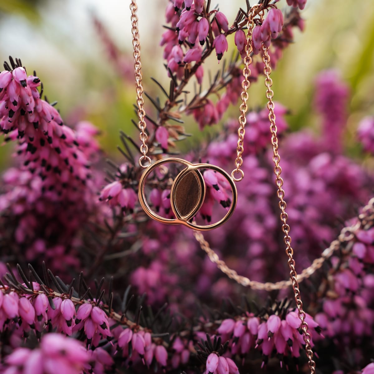 "Effigy Necklace", a stylish necklace in walnut and rose gold, elegantly displayed among vibrant pink flowers.