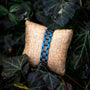 "Triad" bracelet, featuring a maple and blue design, styled elegantly on a rustic cushion surrounded by foliage.