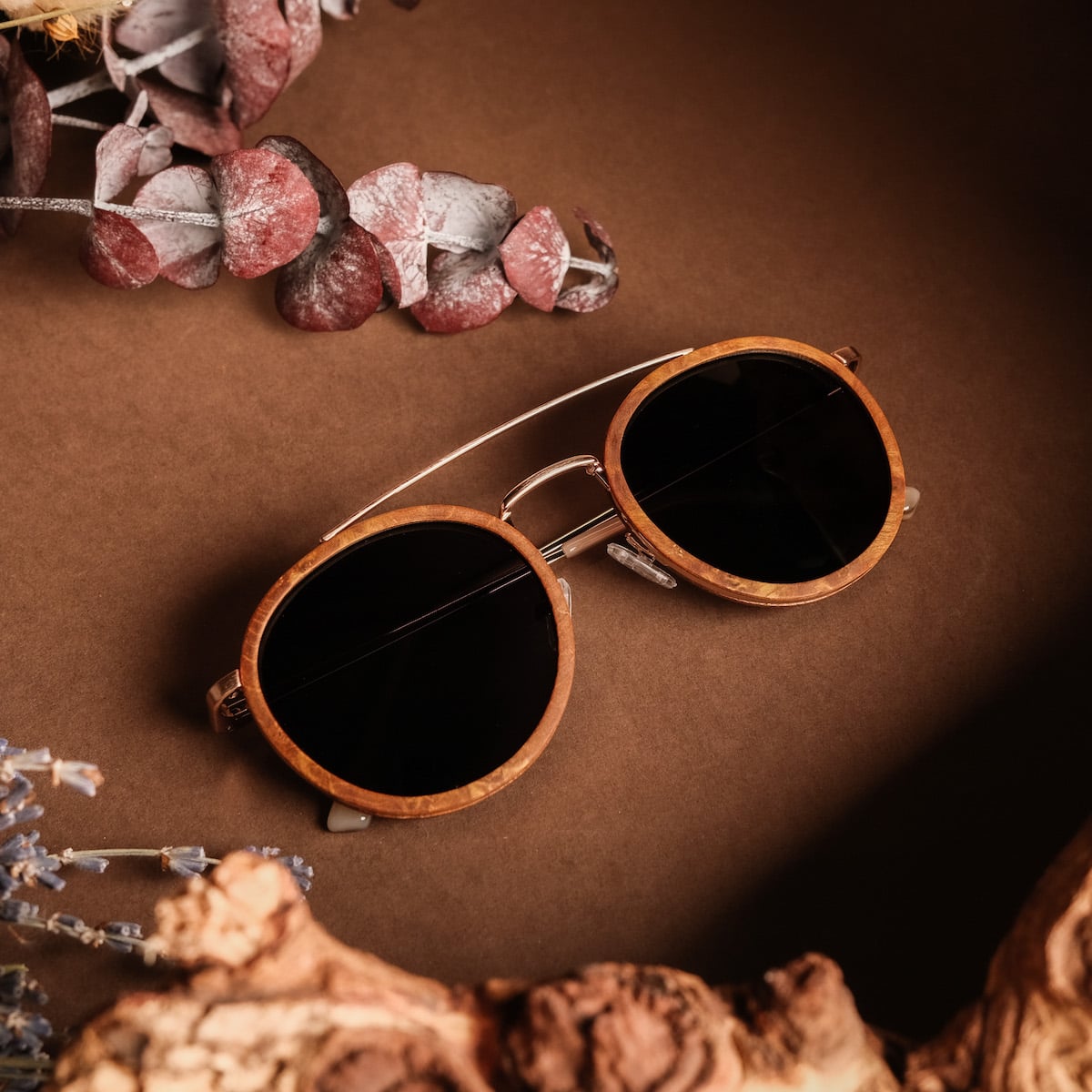 "The Enchanting" sunglasses featuring a chic cinnamon wood frame and matte gold accents, styled elegantly on a natural backdrop.