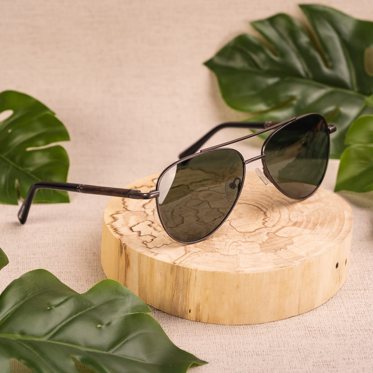 "The Badass II" sunglasses featuring a Leadwood and Antique Silver design on a natural wood display with tropical leaves.