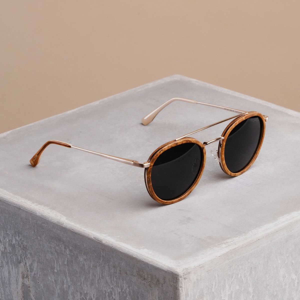 "The Enchanting" sunglasses featuring a cinnamon wood frame and matte gold accents on a minimalist concrete background.