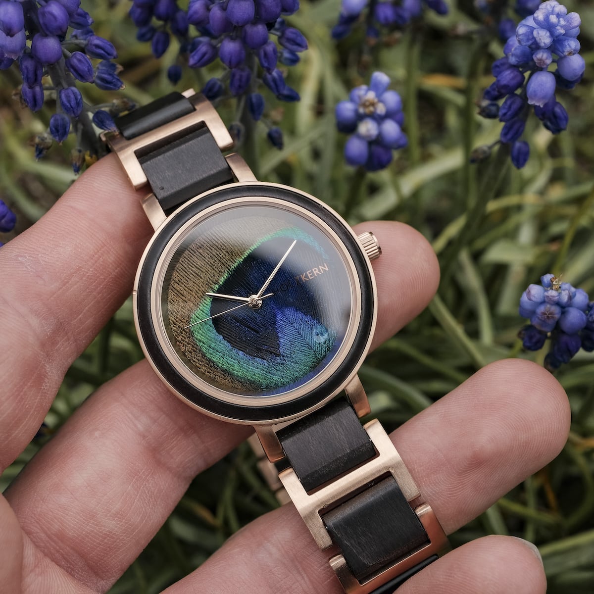 "The Fearless" watch with a striking peacock feather dial and maple elements, set against a backdrop of vibrant flowers.