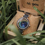 "The Fearless" watch with a maple band and peacock feather face, set in a stylish wooden box surrounded by grass.