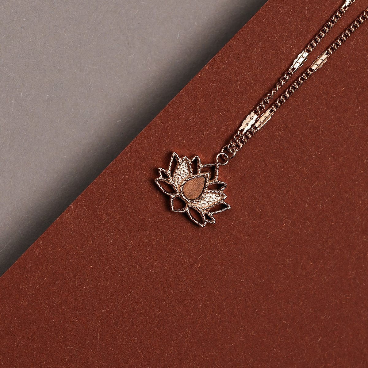 "Balance Necklace", a walnut and rose gold necklace featuring a lotus pendant, styled on a brown and gray background.
