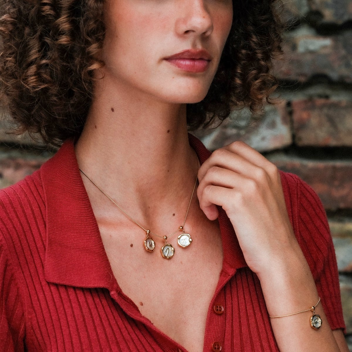 "Letter Charm A-Z", letter charm necklace in nacre and gold, worn by a person in a red top against a brick background.
