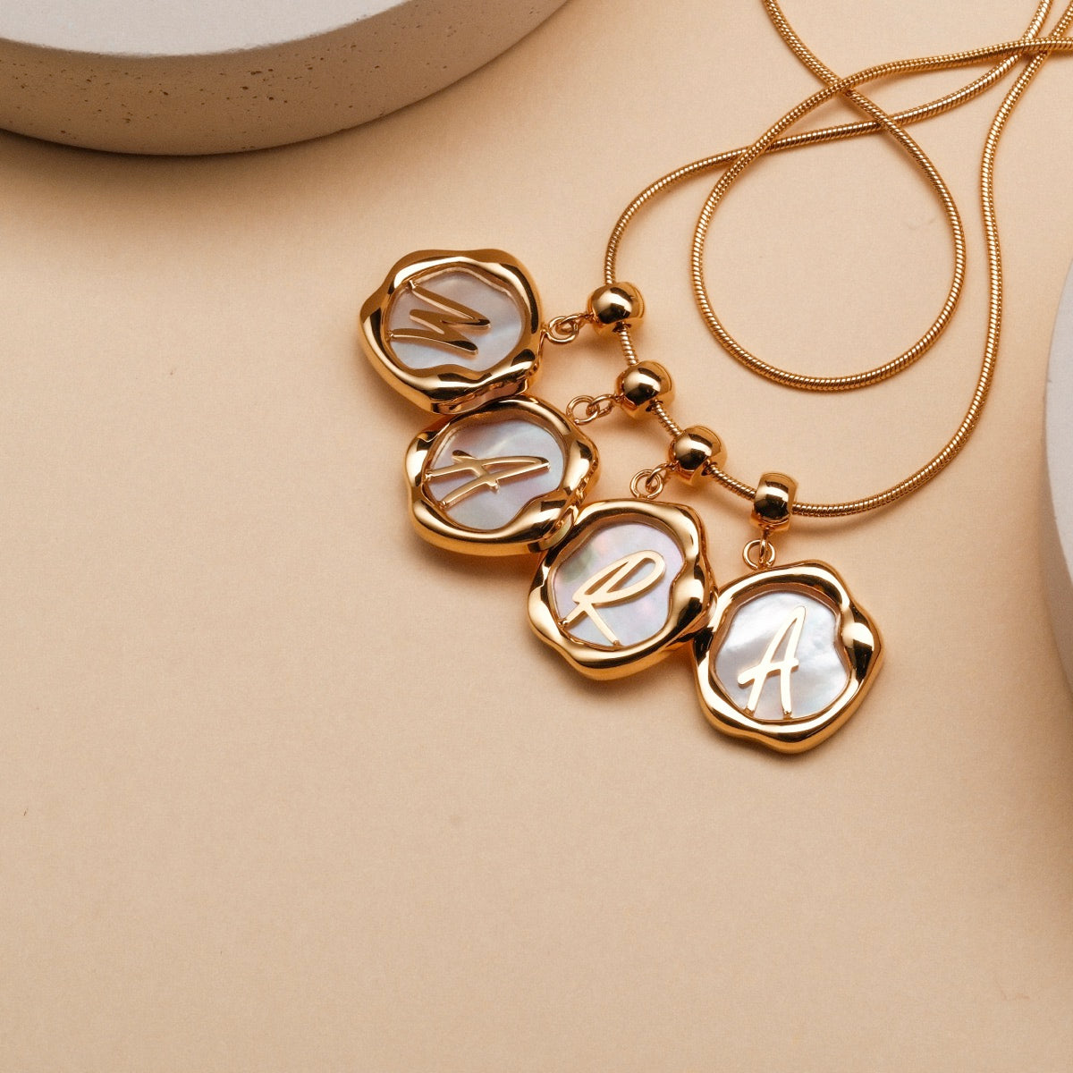 "Letter Charm A-Z" letter charms featuring nacre with gold accents, displayed on a delicate chain.