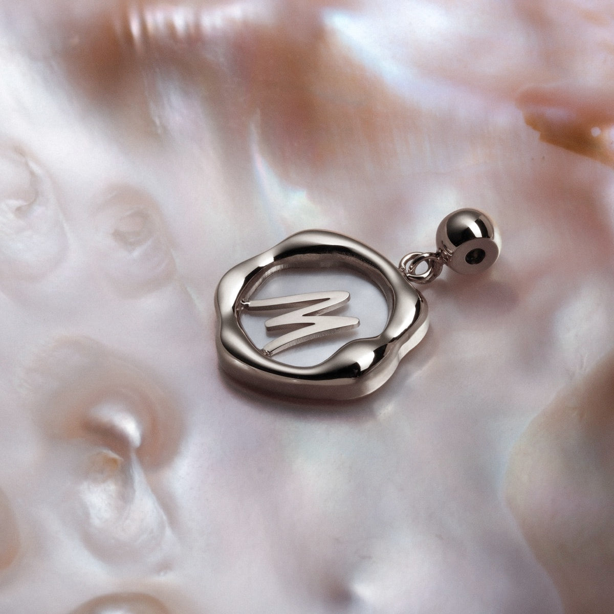 "Letter Charm A-Z" features a sleek silver charm on a nacre background, showcasing elegant letter design in a modern style.