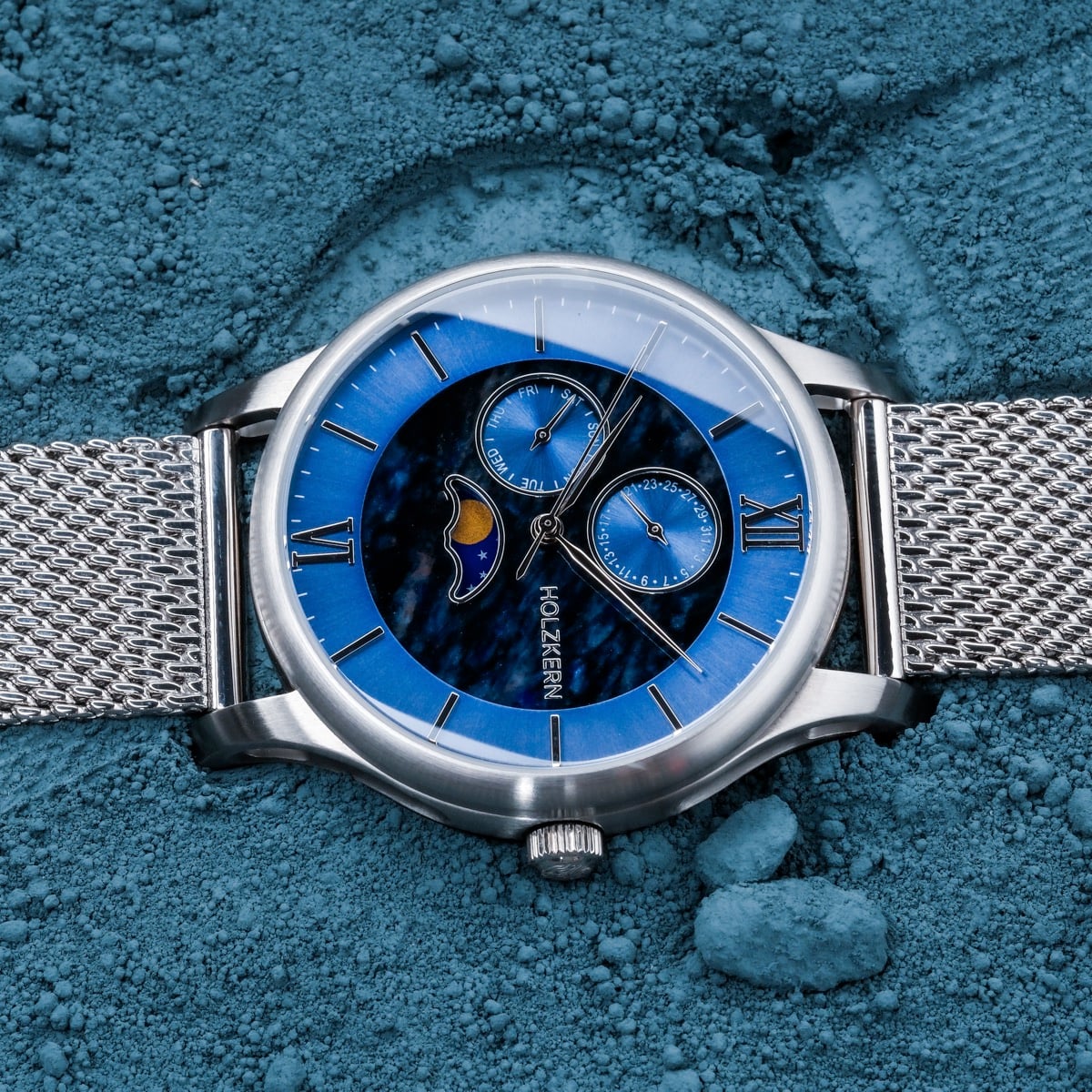 "Moonrise" watch with a marble face, silver accents, and a metal mesh band on a textured blue background.