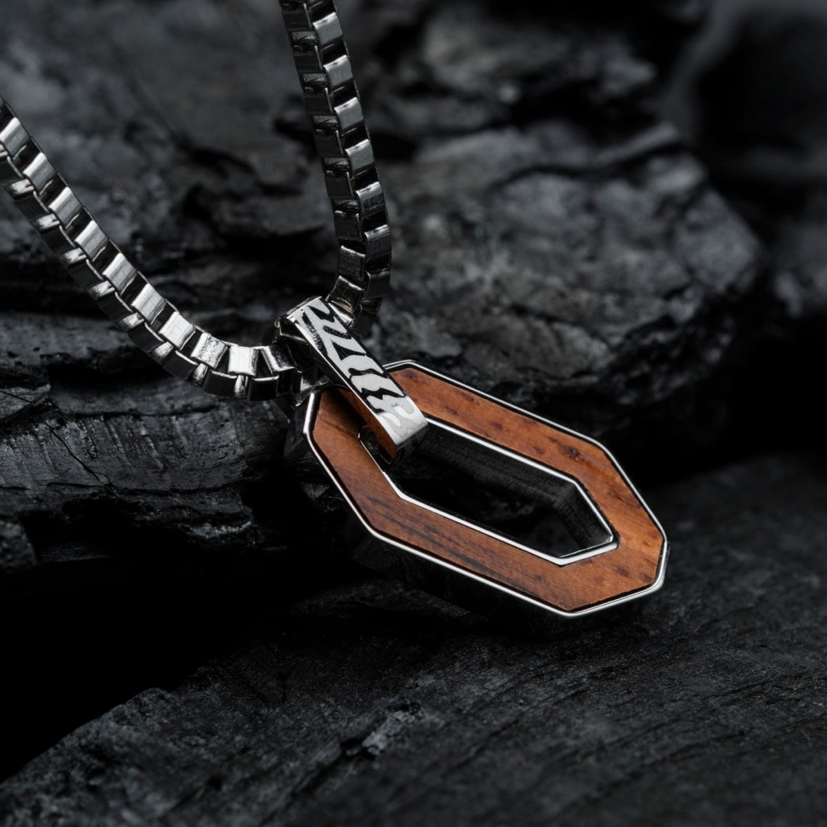 "Interplay Necklace" necklace with a walnut and Damascus design on a sleek metal chain, set against a rugged dark background.