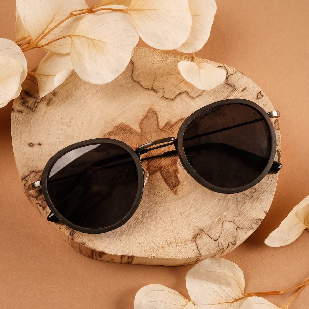 "The Radiant S" sunglasses in Leadwood/Dark Grey, featuring a stylish and natural aesthetic on a circular wooden platform.