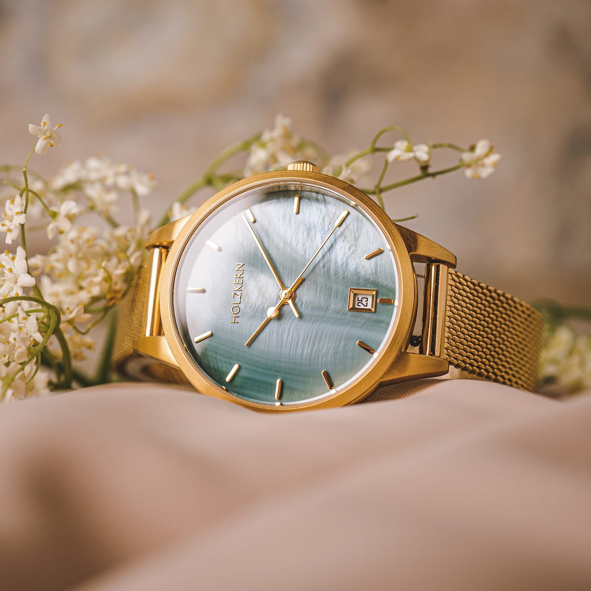 "Talamanca" watch featuring a nacre dial and gold mesh band, surrounded by delicate flowers for an elegant look.