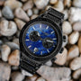 "Aoraki" watch featuring a Lapis Lazuli face and black casing, set against a textured pebble background.