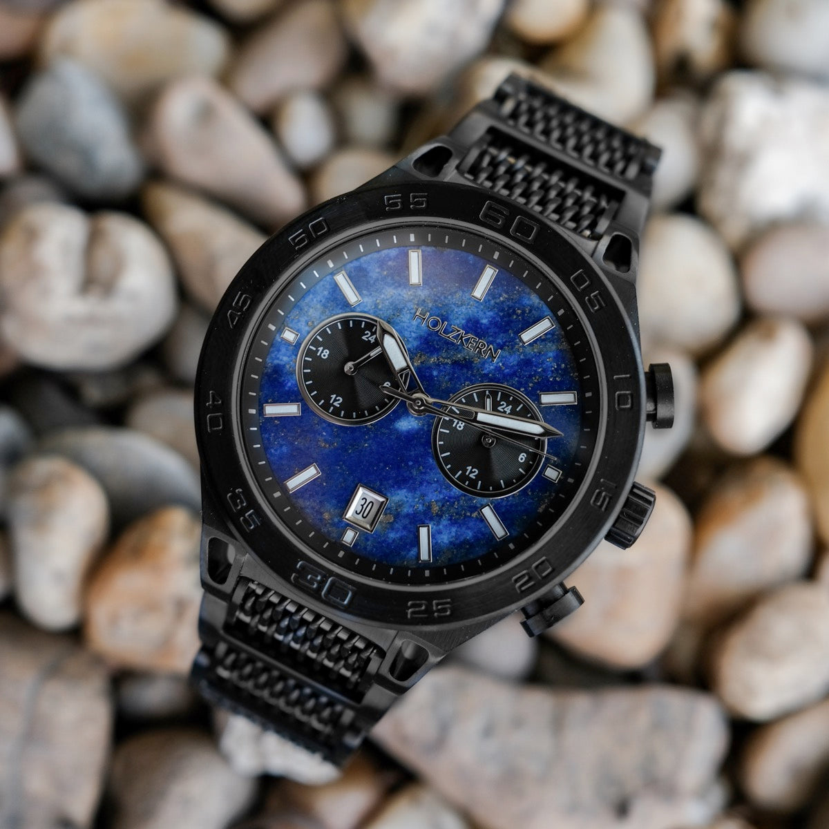 "Aoraki" watch featuring a Lapis Lazuli face and black casing, set against a textured pebble background.