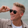 "The Honorable", stylish sunglasses made of leadwood and silver, worn by a smiling person near the water.