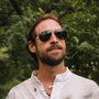 "The Badass II" sunglasses, featuring a Leadwood and Antique Silver design, worn outdoors against a lush green backdrop.
