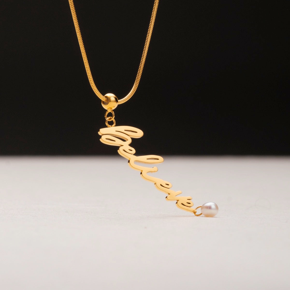 "Believe-Charm", a gold charm necklace featuring a pearl accent, elegantly cursive design.