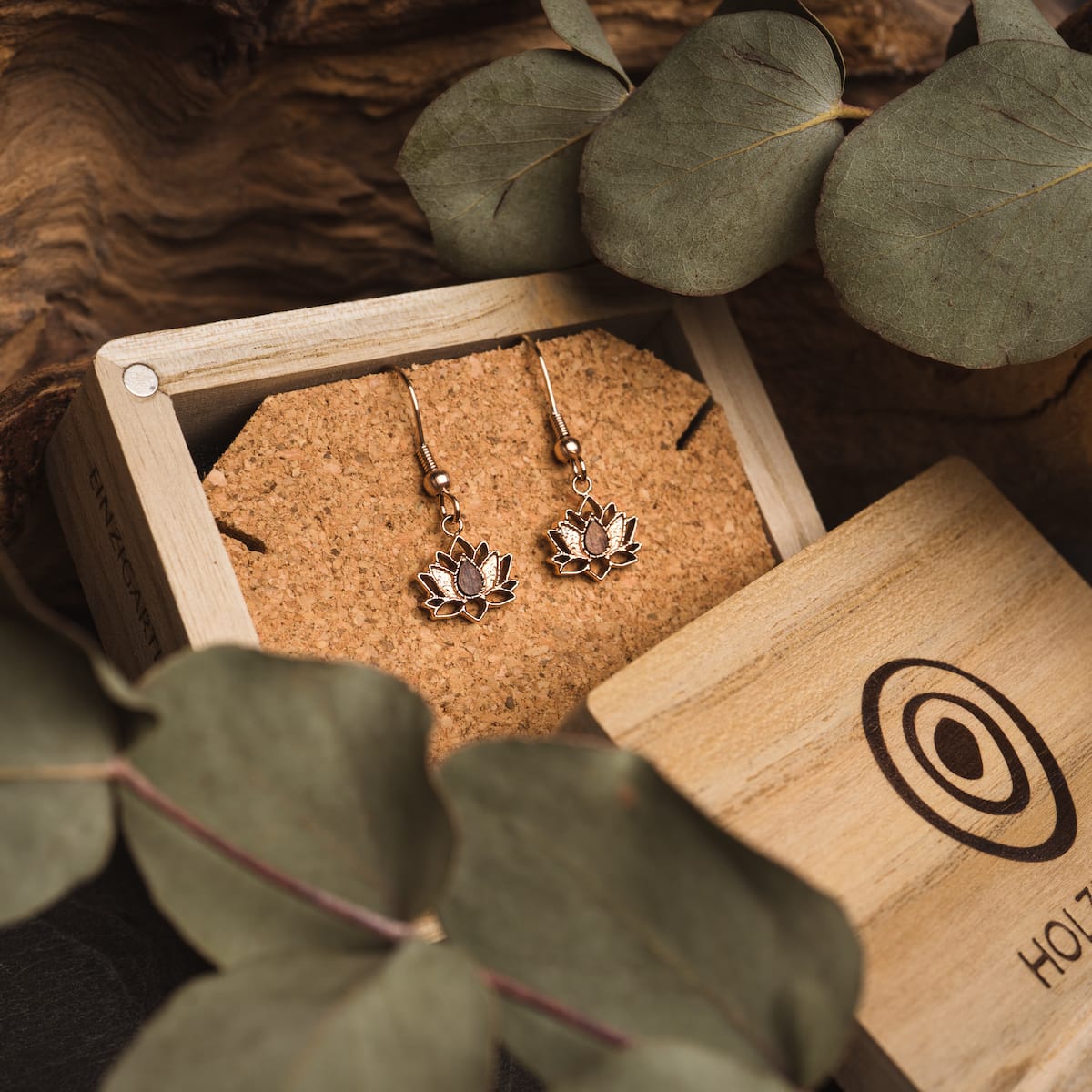 "Balance Earrings", elegant earrings featuring walnut and rose gold lotus design, displayed in a wooden box with green foliage.