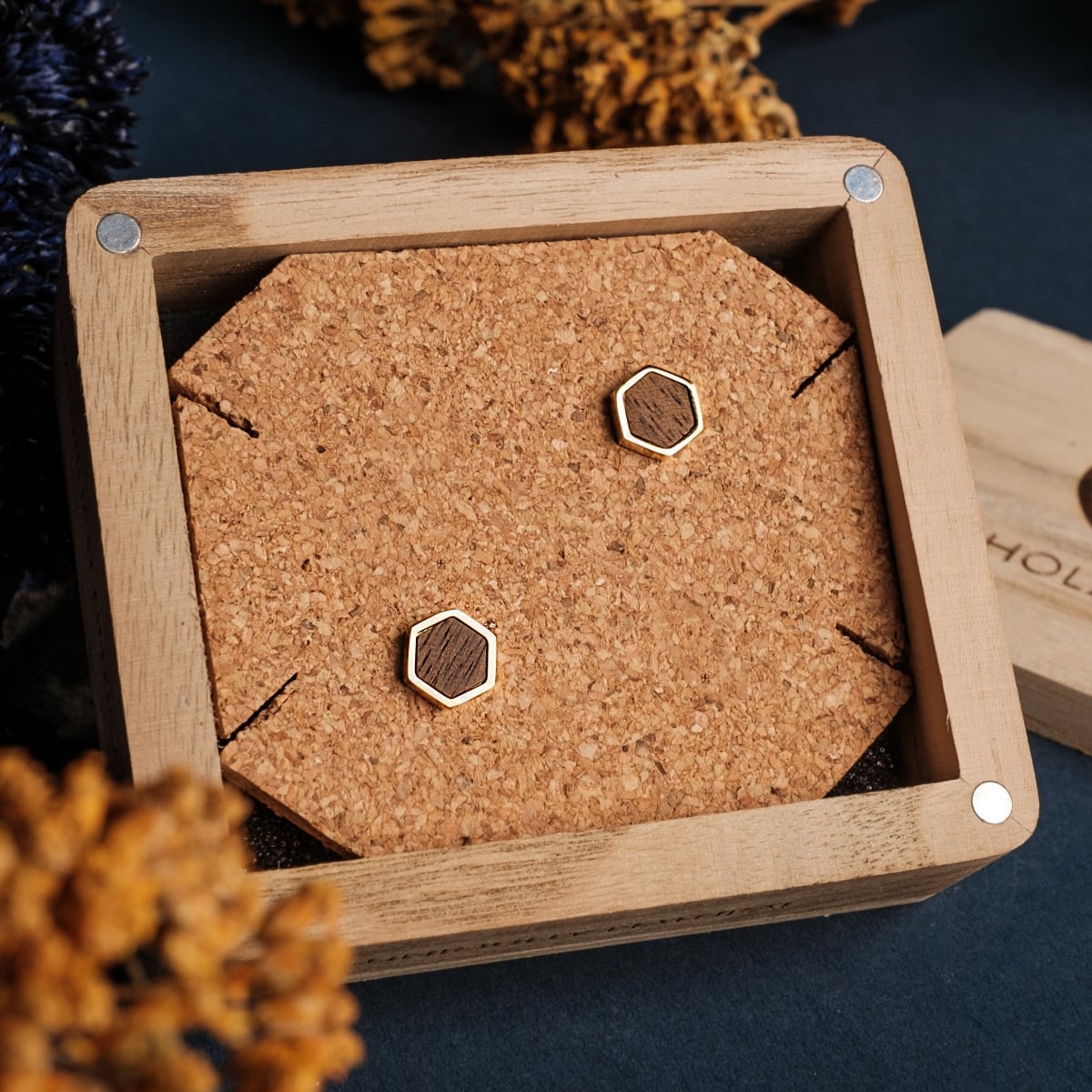 "Facade Earrings" earrings feature a hexagonal walnut and gold design, elegantly displayed on a cork background.