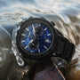 "Aoraki" watch with lapis lazuli dial and black accents, featuring a sleek chronograph design in a dynamic water setting.