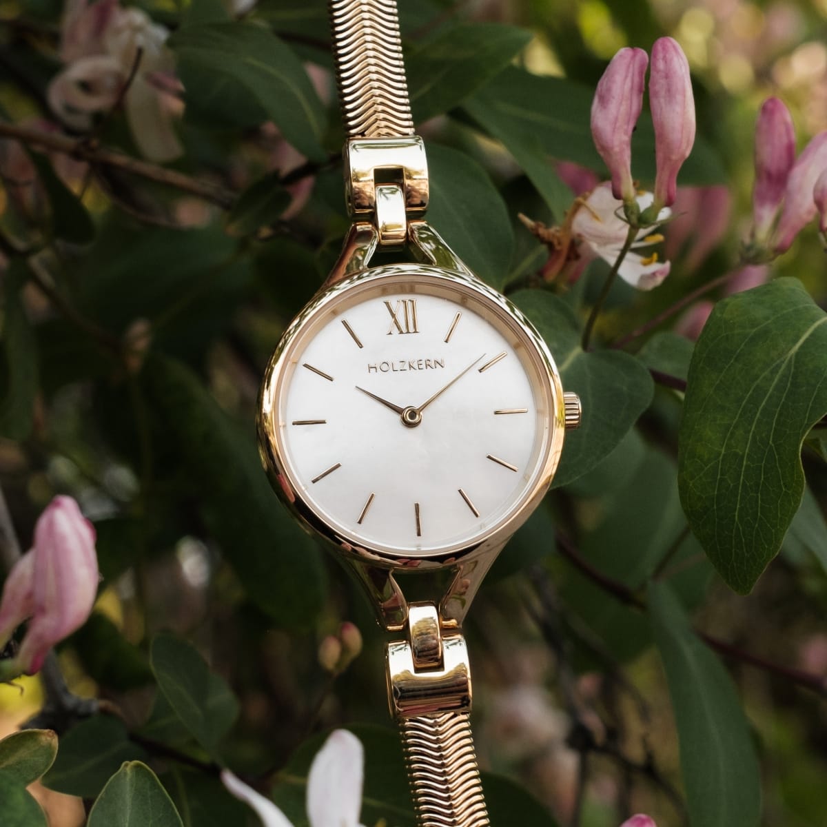 "Kaliya" watch, nacre and gold, elegant design with minimalist style amidst greenery and pink flowers.
