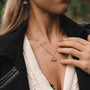"Balance Necklace", an elegant necklace in a gold finish with an olive accent, worn by a person outdoors.
