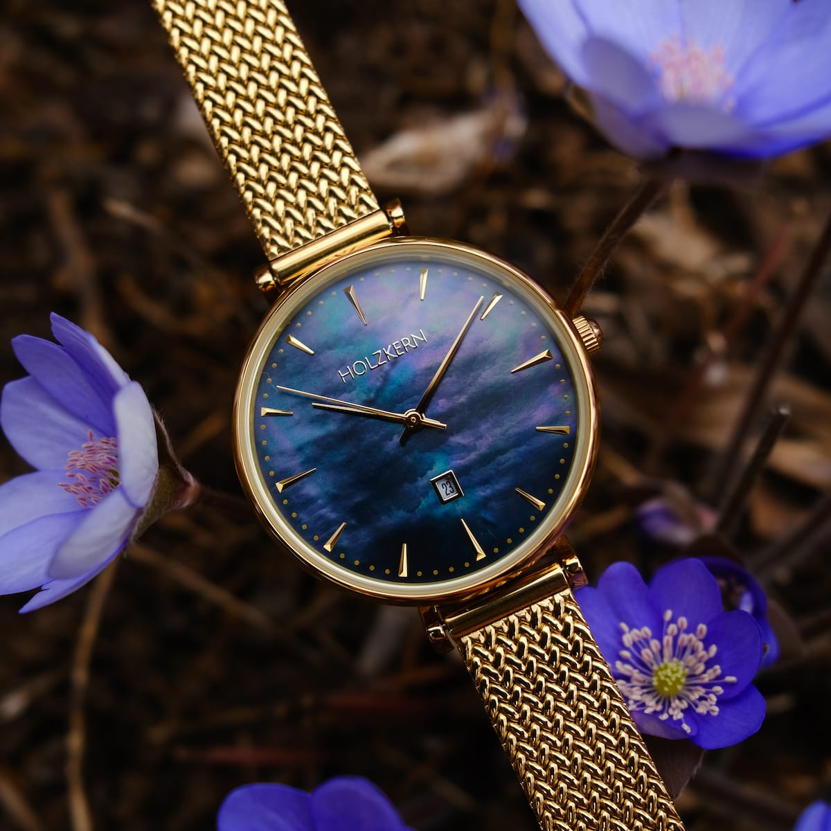 "Giggles" watch with nacre dial and gold mesh band, surrounded by purple flowers.
