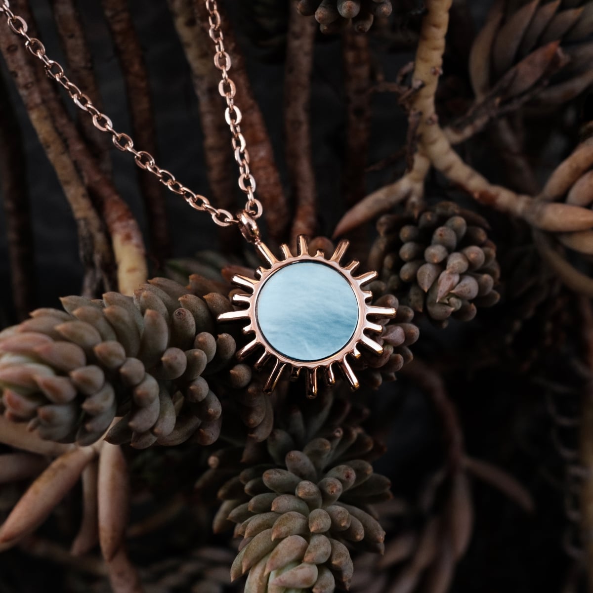 "Shade Necklace", a necklace with a sunburst pendant featuring blue nacre and rose gold, styled against a natural background.