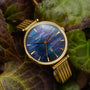 "Giggles" watch with nacre dial and gold accents, showcasing elegant design amidst green leaves.