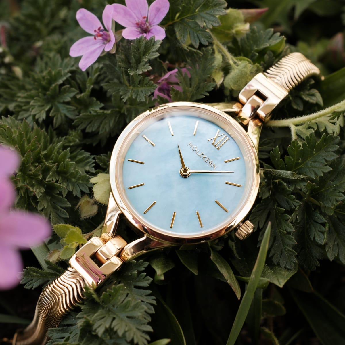 "Shesha" watch with nacre dial and gold accents, showcasing an elegant and sophisticated design amidst green foliage and purple flowers.