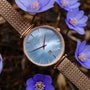 "Chitchat" watch, featuring a nacre dial and rose gold mesh strap, styled among delicate blue flowers.