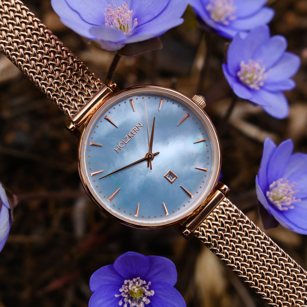 "Chitchat" watch, featuring a nacre dial and rose gold mesh strap, styled among delicate blue flowers.