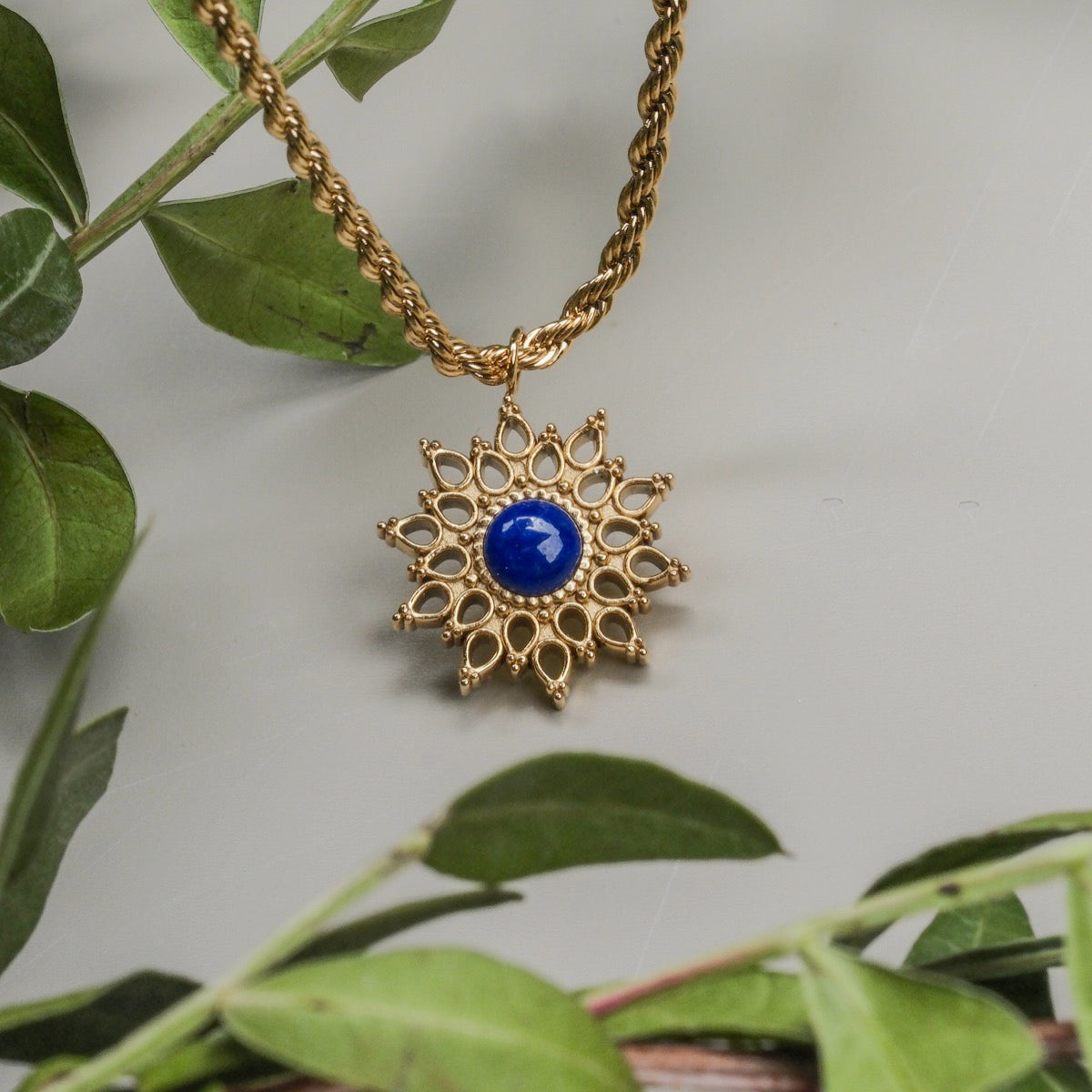 A "Suncatcher Necklace" pendant necklace featuring a sunburst design with lapis lazuli and gold accents, surrounded by green leaves.