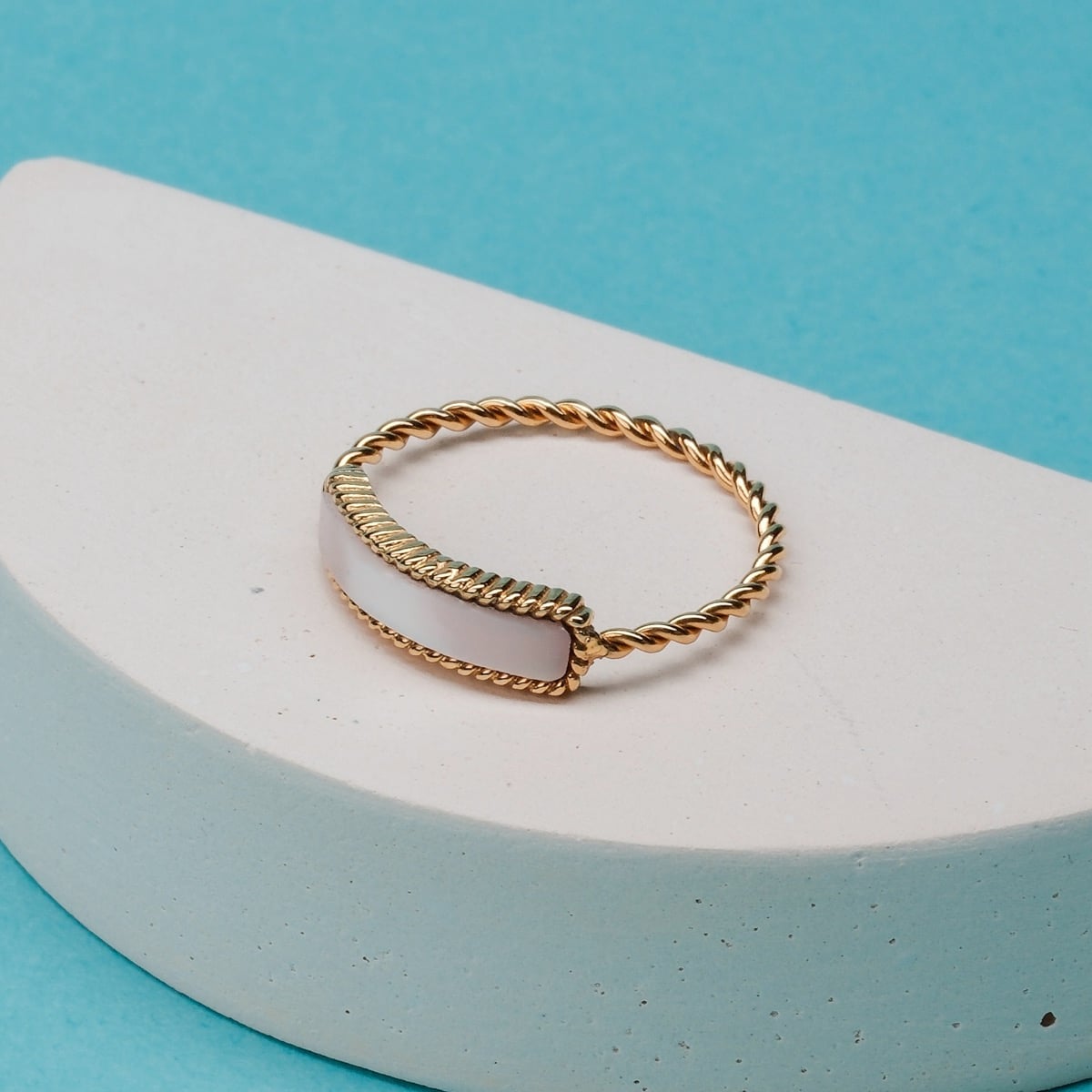 "Summersweet Ring", a gold ring featuring pink nacre, elegantly twisted design against a vibrant blue backdrop.