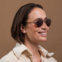 "The Badass II" sunglasses with a walnut and matte gold design, worn by a smiling person.