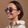 "The Visionary" sunglasses featuring a walnut and bronze style worn by a smiling person in a cozy cream sweater.