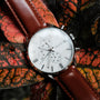 "Melodical" watch with a marble face, silver accents, and brown leather strap on textured leafy background.