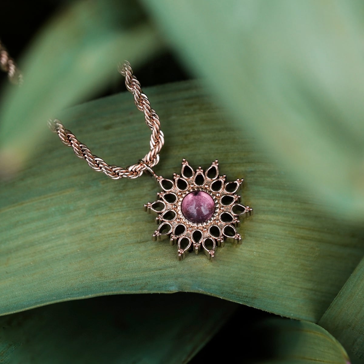 "Suncatcher Necklace", a rose gold suncatcher necklace featuring an amethyst centerpiece, displayed against green foliage.