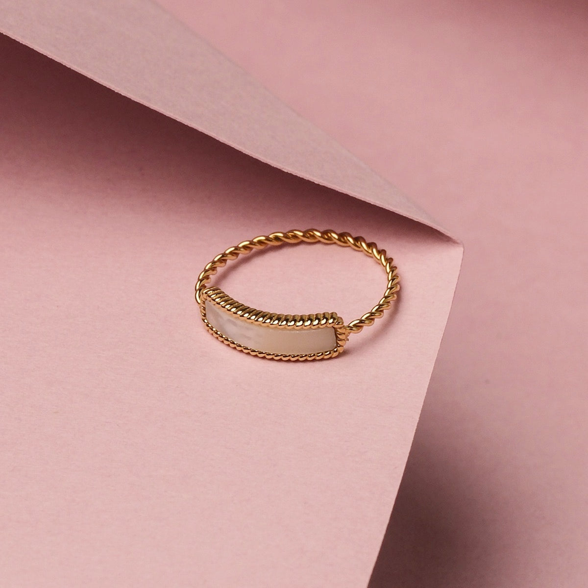 "Summersweet Ring", gold ring with white nacre insert, featuring a twisted band design against a soft pink background.