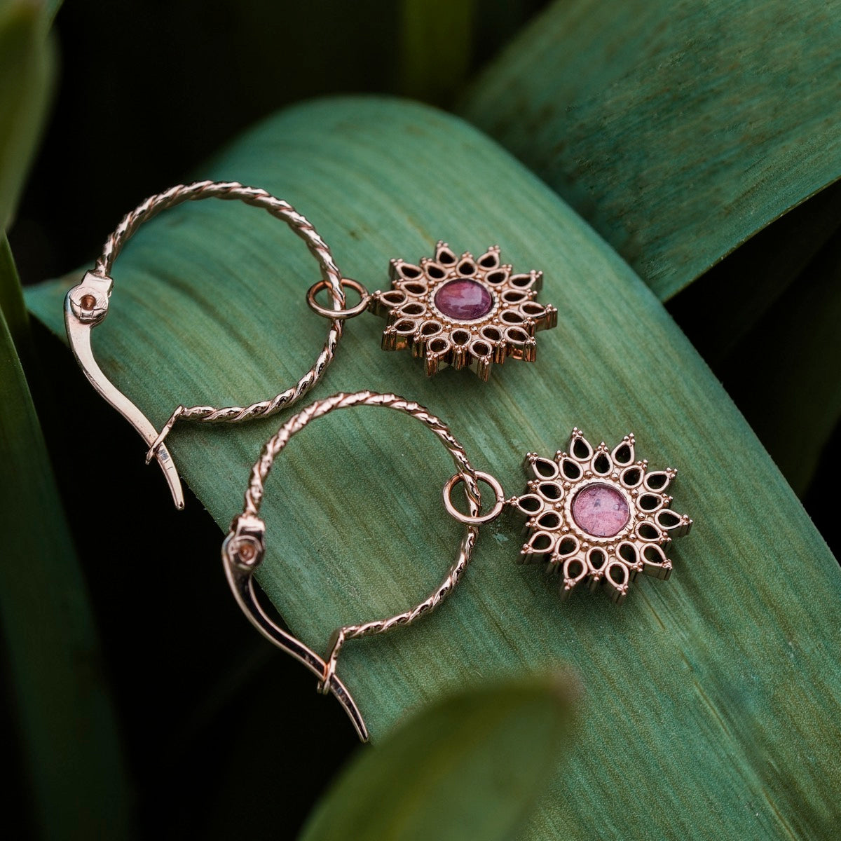 "Suncatcher Earrings", elegant rose gold earrings with amethyst accents, featuring a sunburst design on a lush green leaf background.
