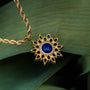 "Suncatcher Necklace", a gold necklace with a lapis lazuli pendant, set against verdant leaves for an elegant contrast.