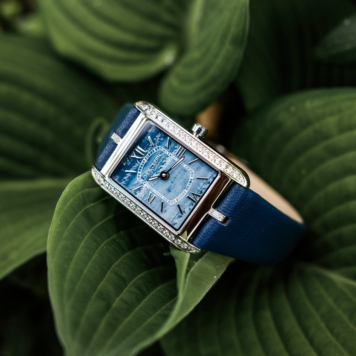 "Memoire" wristwatch features an elegant blue agate face and a refined band, set against lush green leaves.