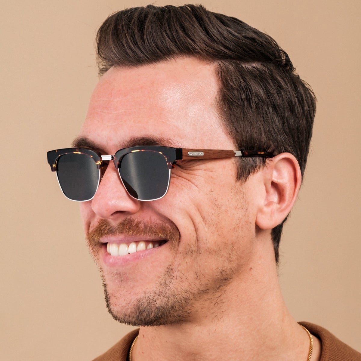 "The Virtuoso" sunglasses featuring a walnut and Havana design, worn by a person smiling against a neutral background.