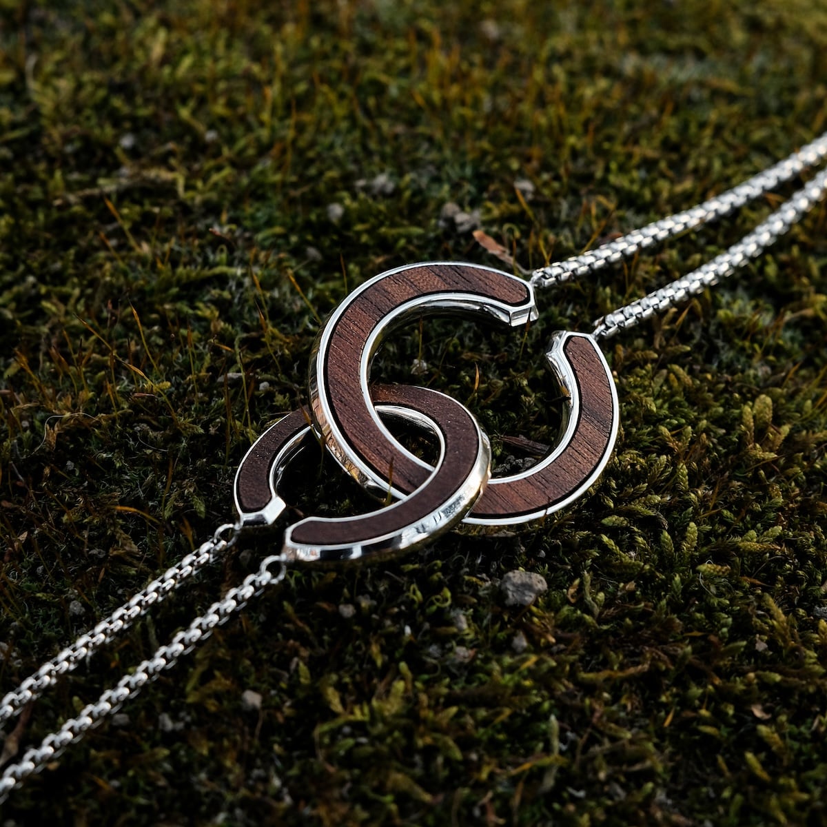 "Inseparable L" necklace featuring intertwined walnut and silver circles on a backdrop of textured moss.