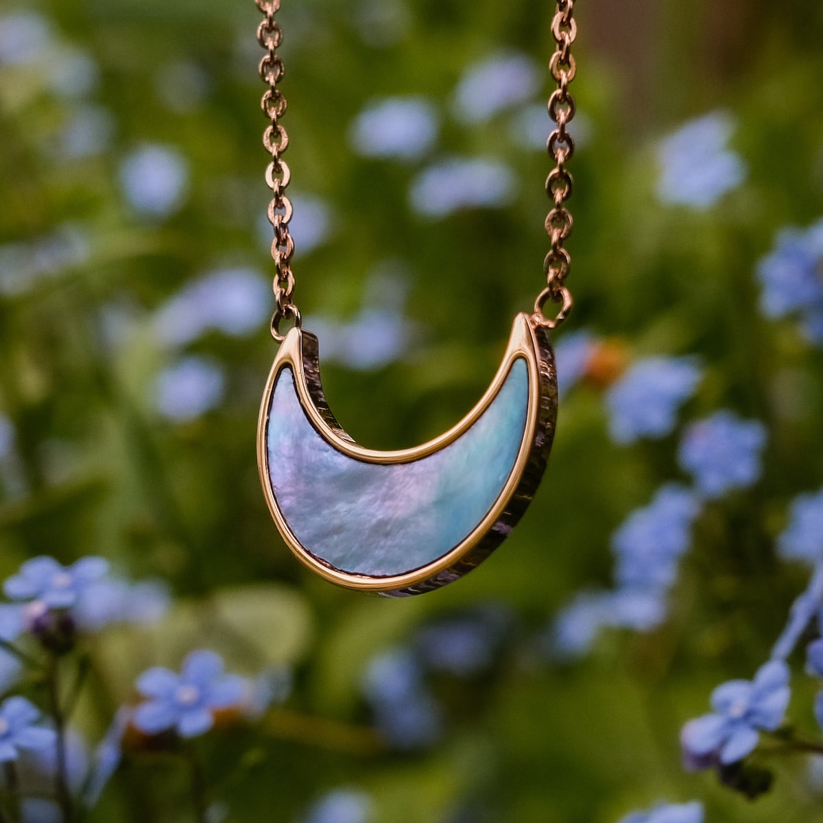 "Hue Necklace", a necklace with blue nacre and rose gold, featuring a crescent moon design against a floral background.