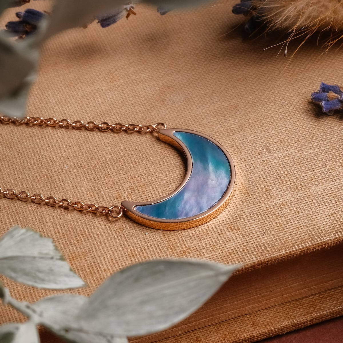 "Hue Necklace" necklace featuring a crescent design with blue nacre and rose gold finish, set against a textured background.